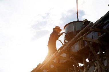 Gün batımında inşaat alanına çimento yükleyen kazıcı ile insan gücü silueti.