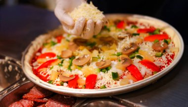 Şefin elinin, taze pizza hamuruna çeşitli malzemelerle mozzarella peyniri serpiştirdiği yakın plan. İtalyan mutfağı, ev yapımı pizza hazırlama ve restoran mutfağı faaliyeti kavramı.