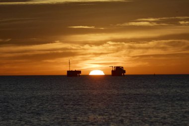 Petrol platformları sakin okyanus ufkunda canlı bir altın gün batımına karşı bir siluet oluşturur.