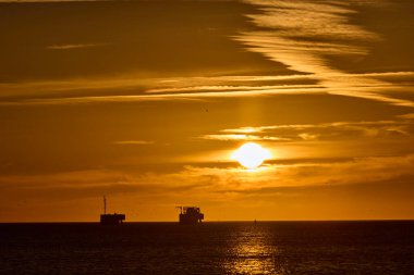 Yanan güneş ufkun altına batıyor, denize ve endüstriyel platformlara turuncu ışık saçıyor.