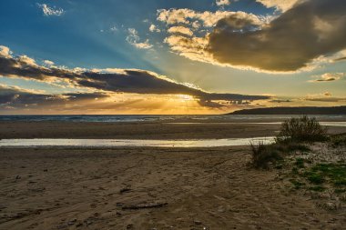 Güneşin batışı bulutların arasından okyanusa akan küçük bir dereyle kumlu bir plajın üzerinden yayılıyor.