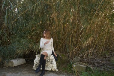 Genç bir kadın yeşil bambu bitkilerinin önünde kayanın üzerinde oturuyor, gülümsüyor ve başka tarafa bakıyor.