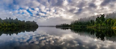 Bulutlar, Yeni Zelanda 'nın batı kıyısındaki Mapourika Gölü' nü çevreleyen bir orman ve çalının sisli sabahını yansıtıyor.