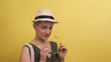 Kameranın önünde gözlüklü ve çantalı bir şapkayla flört eden yetişkin bir kadın. Stüdyoda kısa saçlı sarı arka planda bir kadın..