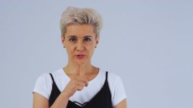 Beyaz arka plandaki yetişkin sarışın kadın stüdyoda kameraya bakıyor. Parmağını dudaklarına koyar..