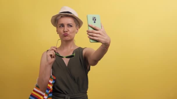 Vuxen modell med flerfärgad halmhatt väska på gul bakgrund. Blond kaukasiska kvinna bär solglasögon och tar selfie. Begreppet resa. — Stockvideo