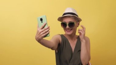 Sarı arka planda çok renkli hasır şapka çantası olan yetişkin bir model. Sarışın beyaz kadın güneş gözlüğü takıyor ve selfie çekiyor. Seyahat kavramı. Boşluğu kopyala.