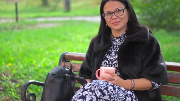 Hermosa mujer asiática en un hermoso vestido se sienta en un parque de otoño en un banco y bebe café de una taza desechable. Mujer madura con café para llevar al aire libre. Imágenes de resolución 4k de alta calidad — Vídeo de stock