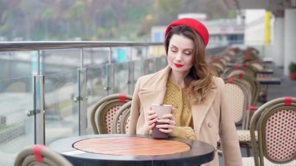 Chica pensativa en una boina roja bebe café caliente o té sentado en la terraza de un restaurante o cafetería en el contexto de una ciudad urbana de otoño. Imágenes de resolución 4k de alta calidad — Vídeo de stock