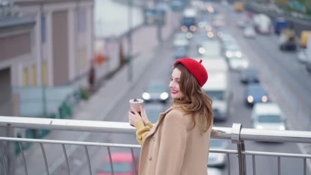 Mujer sonriente joven en boina roja mirar a la ciudad urbana con coches de hora punta de pie en el puente peatonal, hermosa dama al aire libre primer plano lindo cámara lenta. Imágenes de resolución 4k de alta calidad — Vídeo de stock