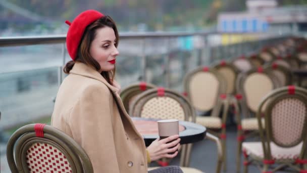 Una joven hermosa en una boina roja bebe café caliente o té sentado en la terraza de un restaurante o cafetería en el contexto de una ciudad urbana de otoño. Imágenes de resolución 4k de alta calidad — Vídeo de stock