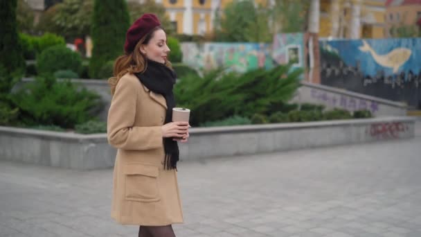 Caminar a una chica encantadora en un abrigo beige y boina roja bebe café al aire libre contra una valla con hermosos graffiti. Imágenes de resolución 4k de alta calidad. 11-11-20 Kiev, Ucrania. — Vídeo de stock