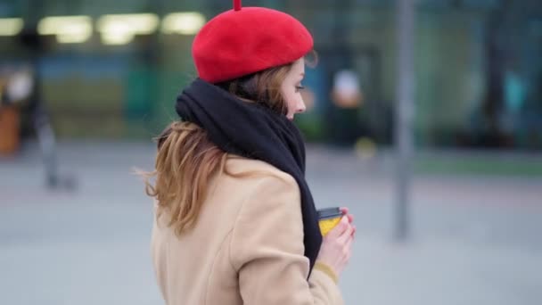 Mujer caminando en la fría ciudad urbana de otoño en boina roja y bufanda negra sosteniendo una taza de café caliente en taza desechable. Chica con estilo disfrutar de la vida urbana entrar en el centro comercial — Vídeo de stock