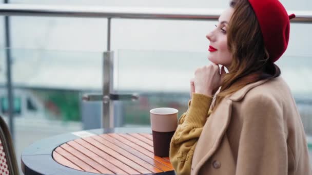 Soñando chica de aspecto francés en una boina roja mirando hacia otro lado mientras bebe café caliente o té sentado en la terraza de un restaurante o cafetería en la ciudad urbana de otoño. 4K UHD — Vídeo de stock