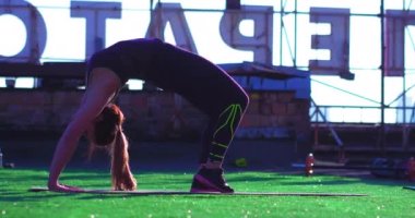 Genç sporcu kız çatıda egzersiz köprüsü yapıyor. Bir ayağı büyük harflerle ve yapay çimlerle evin üzerinde. 10-18-17 Kyiv, Ukrayna. 4k UHD