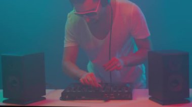 Karıştırma konsolunun gece kulübünde çalışan havalı bir disk jokeyi, gece hayatı kavramının dumanlı siyah arka planında neon renkli ışıklarla görülen kulaklıklarla kulaklıkları dinliyor. Yüksek kalite 4k video