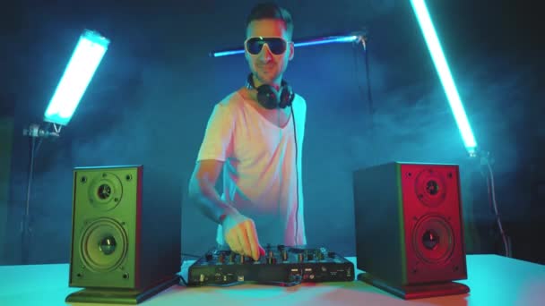 Cool disc jockey actuando en un club nocturno en un controlador de mezclador, visto por luces de colores sobre fondo negro ahumado - vida nocturna concepto 4k metraje — Vídeos de Stock