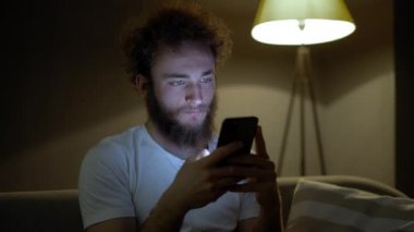 Şirin kıvırcık genç adam internet üzerinden akıllı telefon kullanarak mesaj yazıyor. İnternetteki iş yazışmaları. İnsanlar, teknoloji, yaşam tarzı