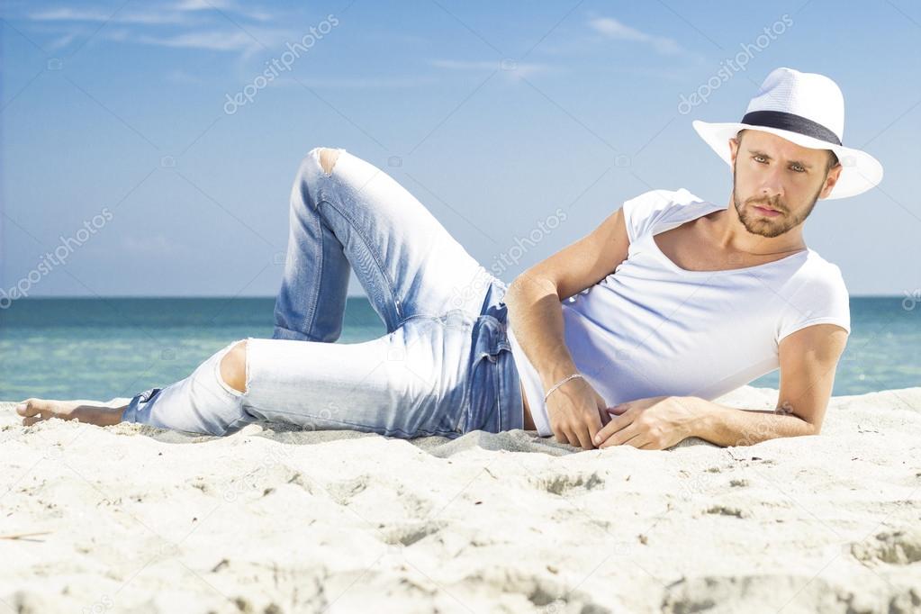 Man on beach lying in sand wearing hipster summer hat. Young male model enjoying summer travel holiday by the sea.