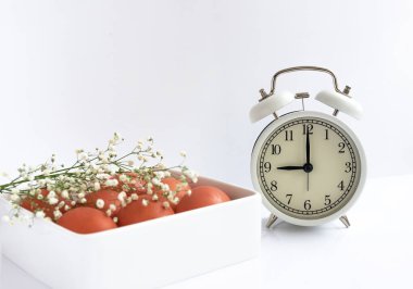 Beyaz kareli tabakta doğal organik yumurtalar ve beyaz bir masada beyaz alarm saati. Yakın çekim, kopyalama alanı. Sağlıklı kahvaltı kavramı, Paskalya