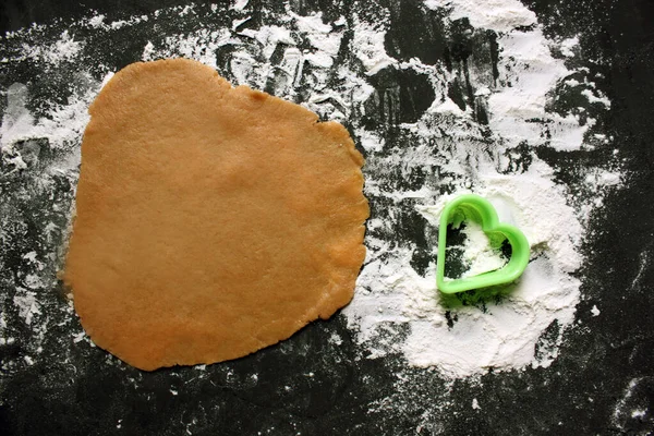 Dough and cookie mold in the shape of heart on black background. Ginger cookie dough on floured black surface. The view from the top. The dough has a brown tint. The concept of cooking. Copy space.