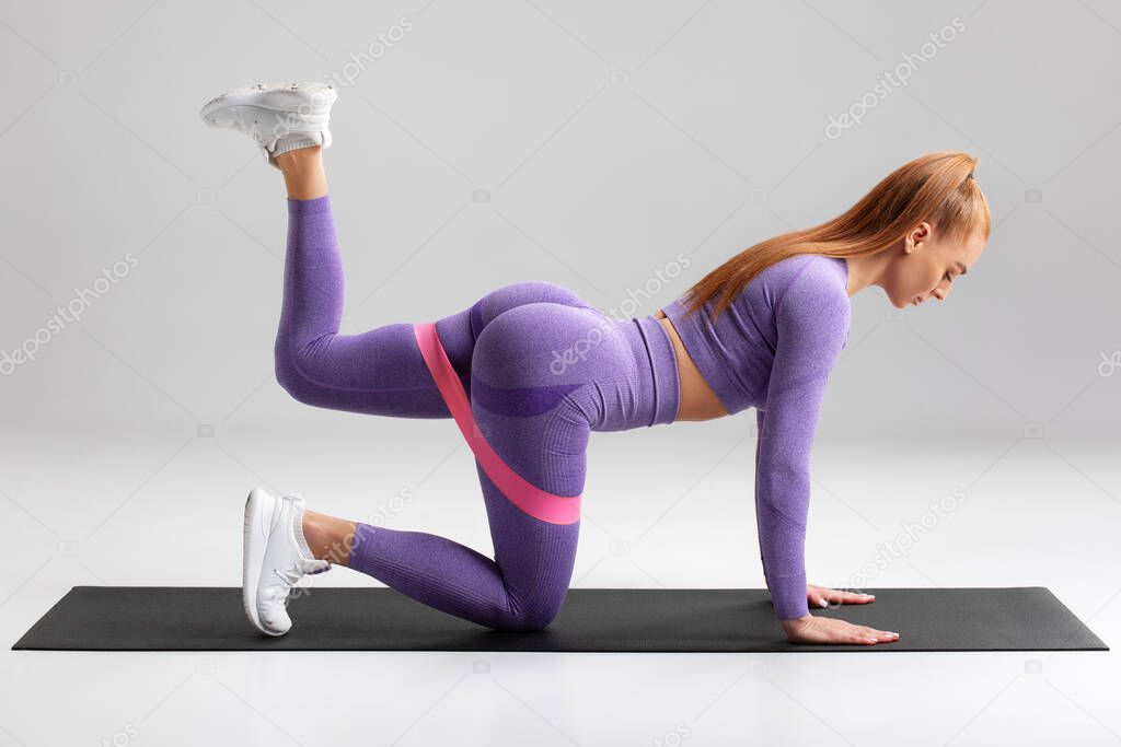 Fitness woman doing kickback exercise for glutes with resistance band on gray background. Athletic girl working out donkey kick