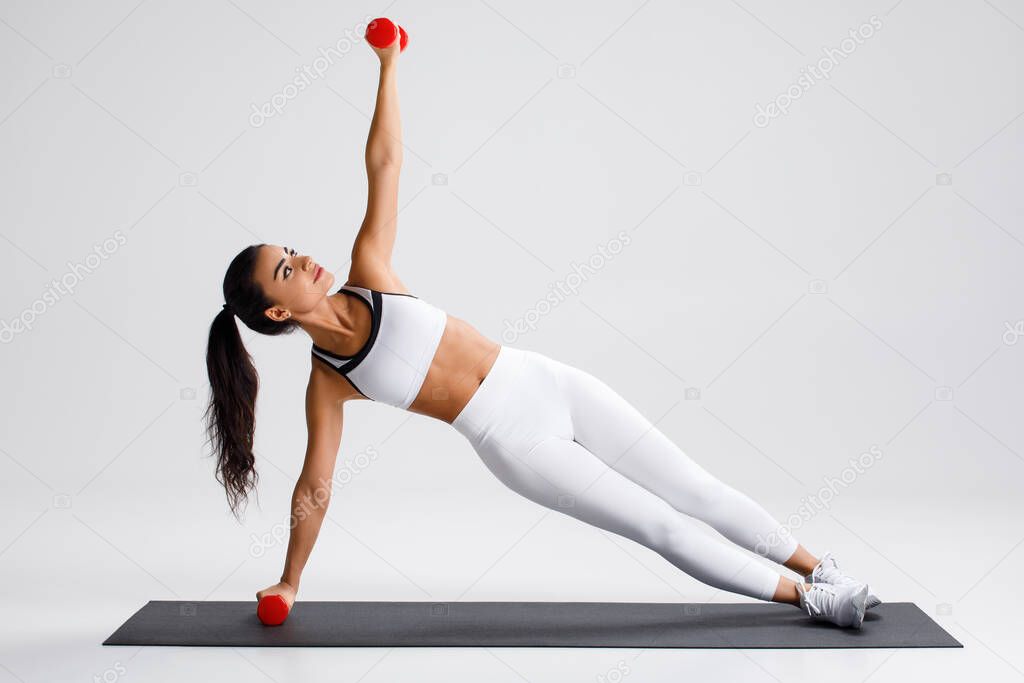 Fitness woman doing side plank exercise on gray background. Athletic girl working out with dumbbells