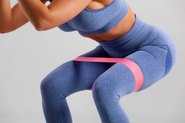 Vücut geliştiren kadın, gri arka planda direnç bandıyla kalça egzersizi yapıyor. Atletik kız spor yapıyor.