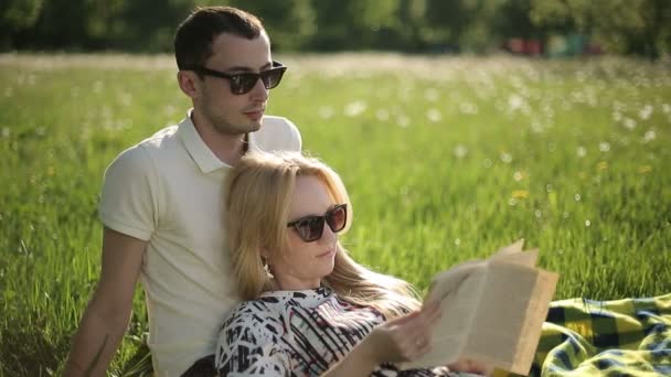 Любящая пара в солнечных очках на закате читает книгу на поле с одуванчиками — стоковое видео