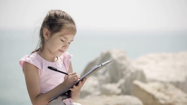 Een kind zit op een steen in de buurt van de Adriatische zee en tekent u een afbeelding. — Stockvideo