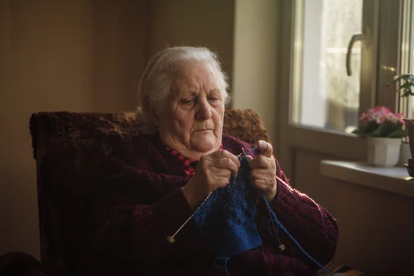 A velha senta-se em casa e tricota roupas — Fotografia de Stock