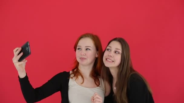 Κορίτσια λαμβάνοντας Αυτοπροσωπογραφία με έξυπνο τηλέφωνο παρακολουθήσετε φωτογραφίες χαμόγελο και το γέλιο. Φόντο κόκκινο στούντιο — Αρχείο Βίντεο
