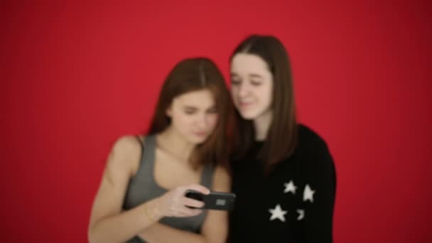 Κορίτσια λαμβάνοντας Αυτοπροσωπογραφία με έξυπνο τηλέφωνο παρακολουθήσετε φωτογραφίες χαμόγελο και το γέλιο. Φόντο κόκκινο στούντιο — Αρχείο Βίντεο