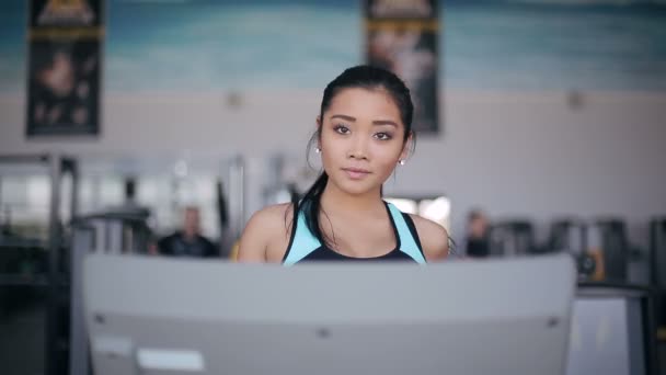 Attraktive asiatische Mädchen läuft auf dem Laufband in der Turnhalle. Porträt mit vollem Gesicht — Stockvideo