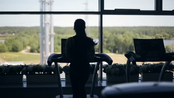 Silhouette eines Mädchens, das auf dem Laufband läuft und in das große Fenster blickt. Schuss von links nach rechts — Stockvideo