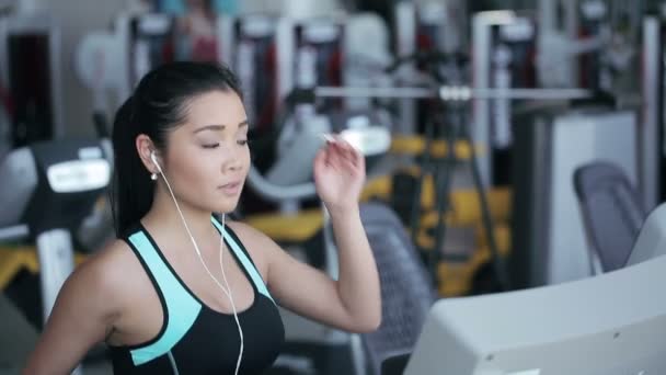 Attraktive asiatische Mädchen, die mit Kopfhörern auf dem Laufband in der Turnhalle läuft. rechtes Profil — Stockvideo