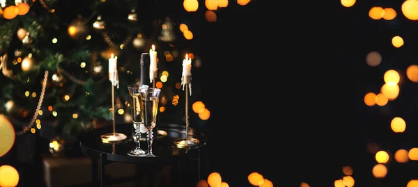 Two glasses and bottle of champagne standing on table with Christmas tree on dark background.