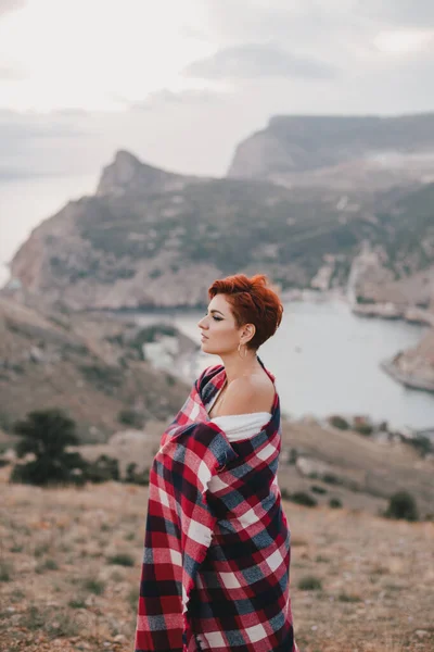 Young woman with short haircut wrapped in red blanket enjoying sunset on the top of the mountain.