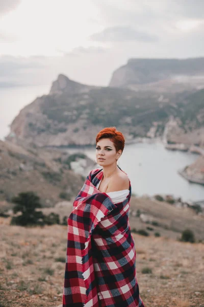 Young woman with short haircut wrapped in red blanket enjoying sunset on the top of the mountain.