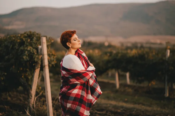 Young woman with short haircut wrapped in red blanket enjoying sunset at the vineyard.