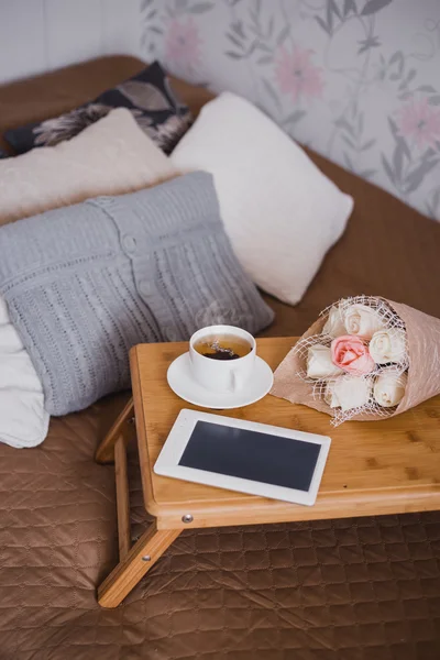 Wooden tray with tea, flowers and e-book standing on a bed