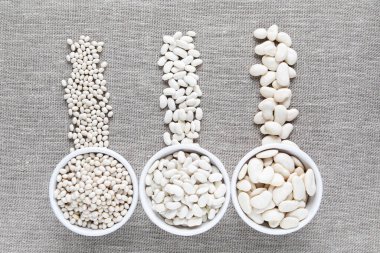 Beyaz kaselerdeki farklı beyaz fasulye türleri ve sağlıklı beslenme ve vejetaryenlik için bez çuval bezi arka planına saçılmış fotokopi alanı ve üst görünümü olan