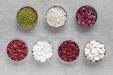 Çeşitli fasulye türleri: keten bezinden yapılmış bir masanın üzerinde farklı boyutlarda renkli fasulyeler - çuval bezi - üst görünüm