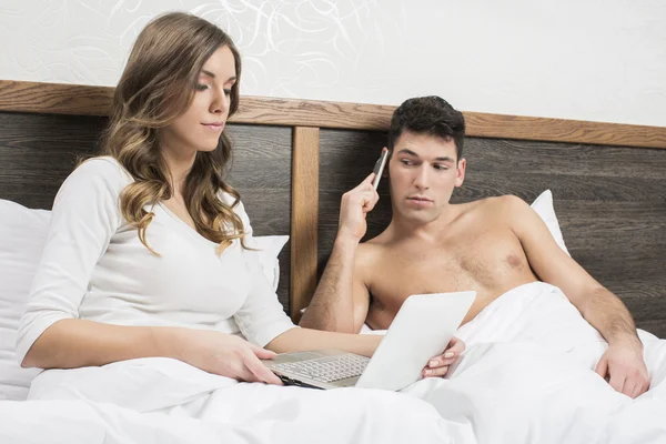 Couple in bed, woman with laptop and man on phone — Stock Photo, Image