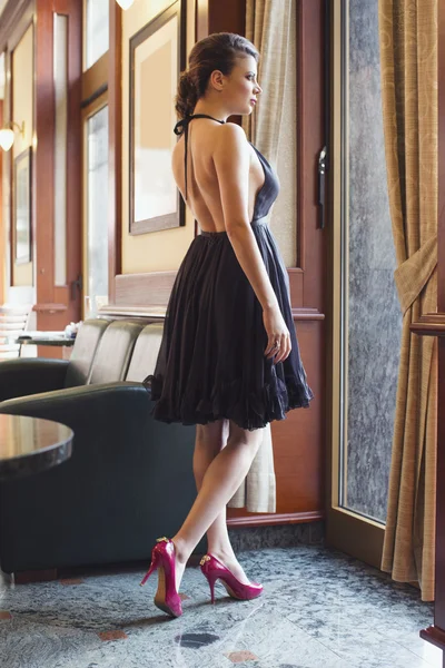 Hermosa joven en vestido negro mirando por la ventana — Foto de Stock