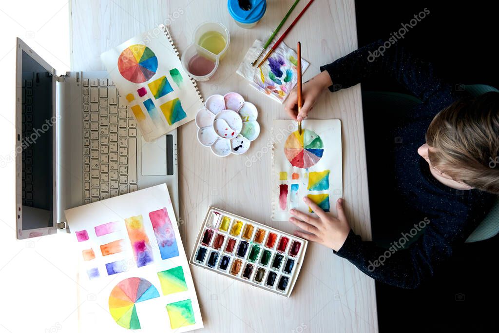 Boy painting pictures with watercolor paints during art lesson. Pupil on drawing with brush. Watercolor color wheel and palette. Color theory beginner hobby lessons