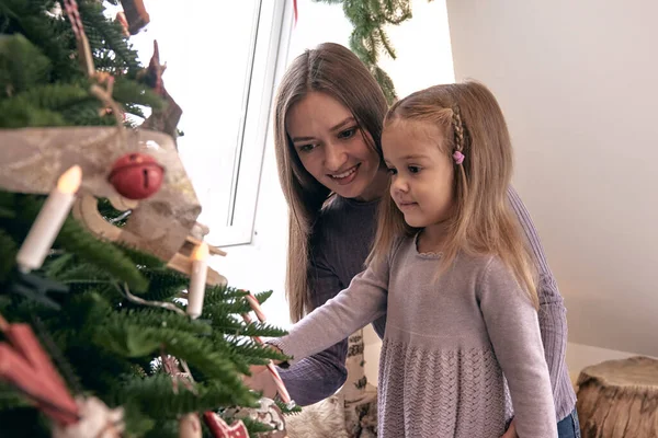 Mom and daughter decorate the Christmas tree. Portrait loving family in Beautiful holdiay DIY handmade home decoration. Cosy room decorated with candles and Christmas tree with presents under it.