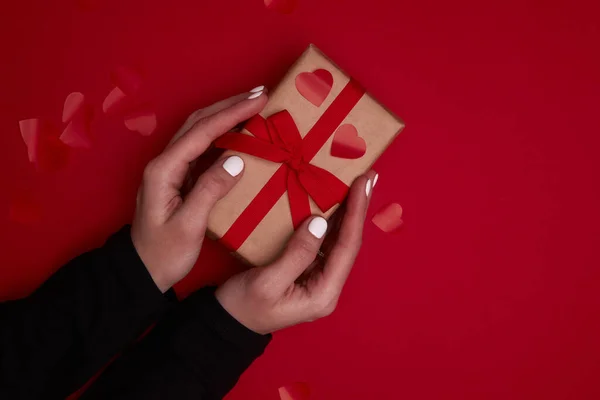 Valentine day composition. Hand holding gift box with ribbon bow and red hearts confetti. — Stock Photo, Image