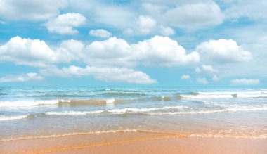 Mavi deniz suyundaki pürüzsüz kumlu kumsal ve beyaz kabarık bulutların altında beyaz dalgalar ve pastel mavi gökyüzü arka planı