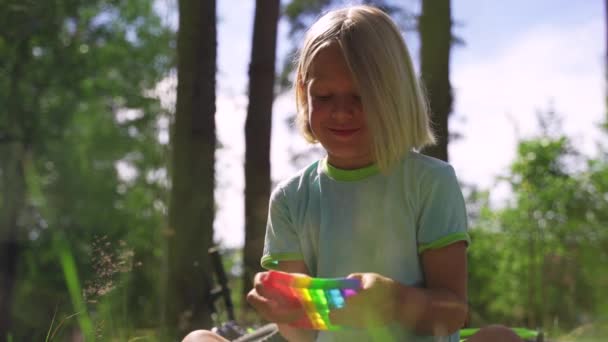 Blondýnka chlapec sedí v parku a hraje popit Rainbow barvy. Stock Video